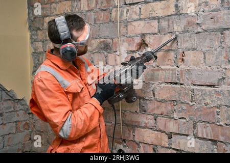 Travailleur de la construction à l'aide d'une perceuse à percussion avec burin pour enlever le vieux ciment du mur de briques à l'intérieur, portant des gants, un masque, des casques antibruit et orange covera Banque D'Images