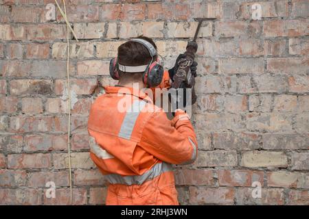 Travailleur de la construction enlevant le vieux plâtre du mur de briques rouges à la perceuse à percussion en position verticale, portant des gants, un masque, un casque antibruit et une couverture orange Banque D'Images