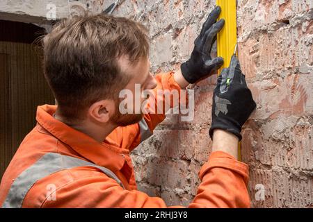 Jeune ouvrier du bâtiment dessinant une ligne verticale sur un mur de briques à l'aide d'un niveau à bulle, portant des gants et une combinaison orange. Banque D'Images