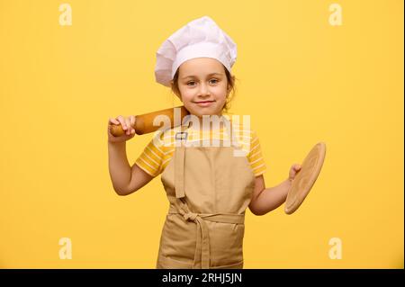 Adorable petite fille, élève de l'école primaire dans le chapeau de chef et tablier, sourit, regardant la caméra, tenant rouleau à pâtisserie et planche en bois, b jaune isolé Banque D'Images