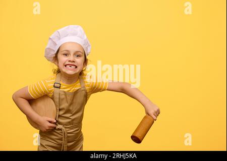 Jeune fille espiègle, élève de l'école primaire en chapeau de chef et tablier, faisant des visages, regardant la caméra, tenant rouleau à pâtisserie et planche en bois, jaune isolé Banque D'Images