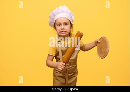 Bel enfant, adorable petite fille caucasienne, élève de l'école primaire portant chapeau de chef et tablier, faisant des visages, regardant la caméra, tenant rollin Banque D'Images