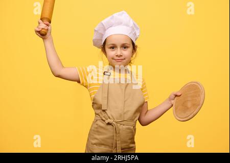 Belle petite fille, élève de l'école primaire en chapeau de chef et tablier, sourit, regardant la caméra, tenant rouleau à pâtisserie et planche en bois, jaune isolé Banque D'Images