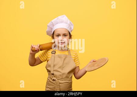 Bel enfant, adorable petite fille caucasienne, élève de l'école primaire portant chapeau de chef et tablier, faisant des visages, regardant la caméra, tenant rollin Banque D'Images