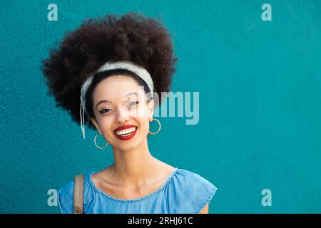 Portrait de jeune femme afro-américaine optimiste avec coiffure afro et boucles d'oreilles avec maquillage souriant et regardant la caméra contre backgroun bleu Banque D'Images