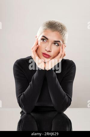 Charmante jeune femme aux cheveux rasés courts portant une chemise noire appuyée sur les mains tout en rêvant et regardant loin dans le studio Banque D'Images