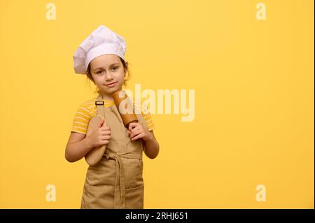 Portrait de studio d'adorable charmante petite pâtissière caucasienne confiseuse en tablier beige et chapeau, tenant une planche en bois et rouleau à pâtisserie, sourires c Banque D'Images