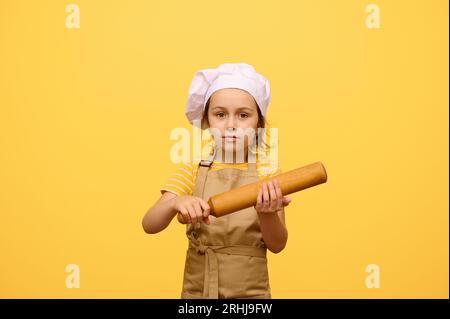 Portrait d'une petite fille en uniforme de chef, tenant un rouleau à pâtisserie en bois, regardant la caméra, fond jaune isolé Banque D'Images