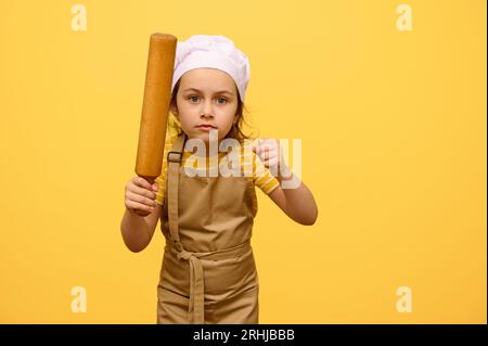 Portrait d'une petite fille en colère sérieuse en uniforme de chef, tenant un rouleau à pâtisserie en bois, serrant les poings, regardant la caméra, exprimant furiosit Banque D'Images