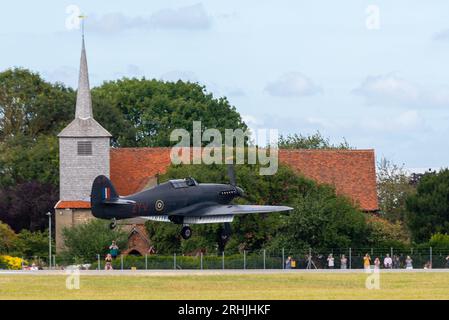 Aéroport de Londres Southend, Essex, Royaume-Uni. 17 août 2023. Les anciens avions Battle of Britain Memorial Flight de la RAF utilisent l’aéroport civil relativement calme d’Essex pour opérer à partir des spectacles aériens en bord de mer de ce week-end à Eastbourne et Folkestone. L’avion de chasse Hurricane est représenté en train d’atterrir, passant devant St Laurence et All Saints Church sur le périmètre de l’aéroport. Les gens sont venus les voir arriver Banque D'Images