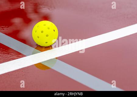 Un pickleball sur un court de tennis de pickleball rouge humide avec des lignes blanches combinées avec des lignes de pickleball grises Banque D'Images