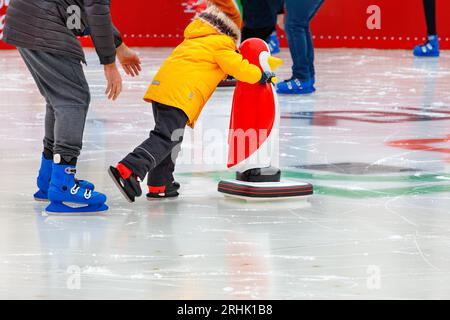 Un enfant, accompagné d'un adulte, apprend à patiner sur une patinoire, se tenant à un assistant, un pingouin jouet. Banque D'Images