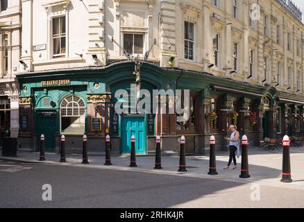 Jeune femme utilisant un téléphone portable passe devant le pub Railway Tavern au coin de Liverpool Street et Old Broad Street. Londres, Angleterre, Royaume-Uni Banque D'Images