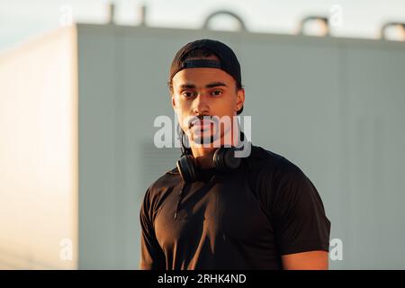 Portrait d'un athlète masculin confiant portant une casquette noire, un casque sans fil et des vêtements de sport regardant l'appareil photo Banque D'Images