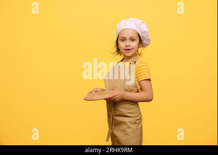 Enfant adorable caucasien, une écolière habillée en chef confiseur, tenant une planche de bois, souriant regardant la caméra, isolée au-dessus de studi jaune Banque D'Images