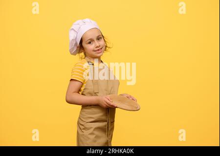 Caucasien mignon petit chef confiseur, cuisinier, boulanger, pizzaiolo, adorable petite fille 6 yeras ol dans le chapeau de chef blanc et tablier de cuisine beige, tenant un W Banque D'Images