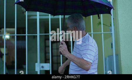 Homme senior ouvrant parapluie pour protéger de la pluie, sort dans la rue de la maison. Personne âgée quittant la résidence, ouvre la porte marche dans le trottoir urbain Banque D'Images