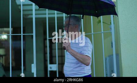 Homme senior ouvrant parapluie pour protéger de la pluie, sort dans la rue de la maison. Personne âgée quittant la résidence, ouvre la porte marche dans le trottoir urbain Banque D'Images