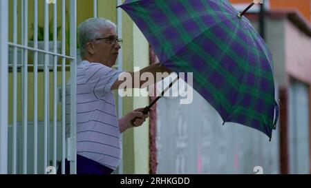 Homme senior ouvrant un parapluie, sort dans la rue sous la pluie, ferme la porte de la maison derrière lui. Personne âgée quittant sa résidence le jour de pluie à Slow-M. Banque D'Images