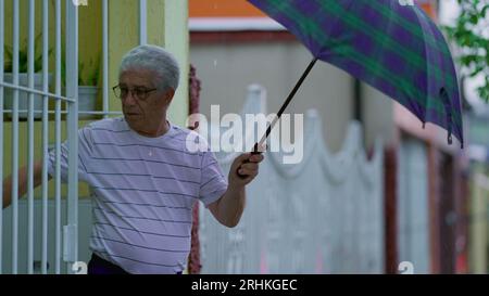 Homme senior ouvrant un parapluie, sort dans la rue sous la pluie, ferme la porte de la maison derrière lui. Personne âgée quittant sa résidence le jour de pluie à Slow-M. Banque D'Images