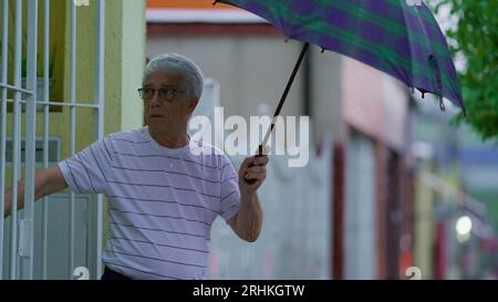 Homme senior ouvrant un parapluie, sort dans la rue sous la pluie, ferme la porte de la maison derrière lui. Personne âgée quittant sa résidence le jour de pluie à Slow-M. Banque D'Images