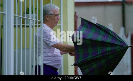 Homme senior ouvrant un parapluie, sort dans la rue sous la pluie, ferme la porte de la maison derrière lui. Personne âgée quittant sa résidence le jour de pluie à Slow-M. Banque D'Images