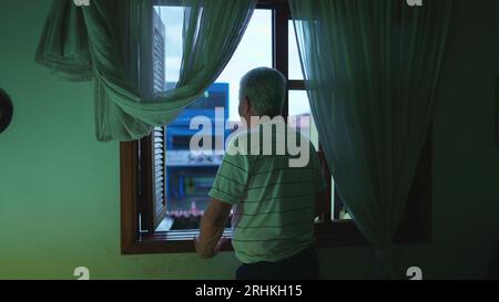 Homme senior debout près de la fenêtre de la chambre dans la soirée regardant la pluie de l'après-midi tomber. Personne âgée contemplative retraitée aux cheveux gris observant neighborho Banque D'Images