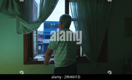 Homme senior debout près de la fenêtre de la chambre dans la soirée regardant la pluie de l'après-midi tomber. Personne âgée contemplative retraitée aux cheveux gris observant neighborho Banque D'Images