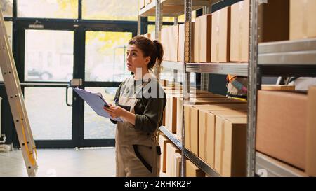 Travailleur de dépôt vérifiant l'inventaire de stock, travaillant sur l'inspection logistique pour expédier les commandes de produits de la salle de stockage. Femme gérant des boîtes de marchandises de fret avec liste pour le contrôle de la qualité. Banque D'Images