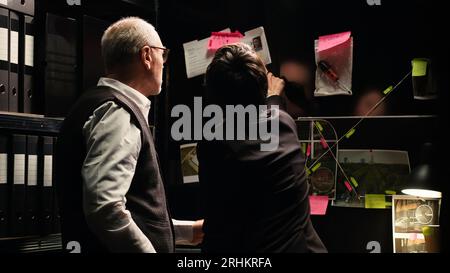 Les agents de police examinent le tableau des preuves dans le bureau d'enquête, analysant les indices découverts pour construire le dossier de l'affaire. Équipe de détectives travaillant avec des documents et des dossiers classifiés pour attraper le suspect. Banque D'Images