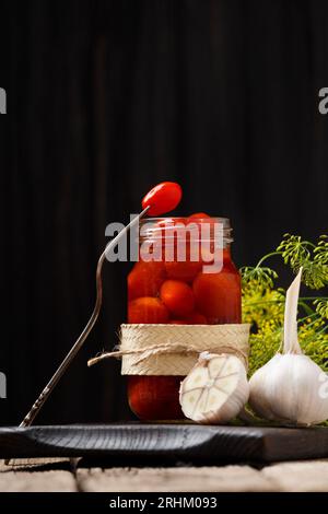 Tomates cerises marinées dans un bocal ouvert sur une planche en bois sombre, une tomate sur une fourchette vintage sur un bocal, fond sombre, gros plan. Banque D'Images