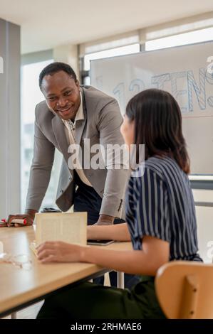 Un professeur d'anglais masculin afro-américain professionnel et joyeux ou tuteur enseigne l'anglais et parle avec une étudiante asiatique pendant le cla Banque D'Images