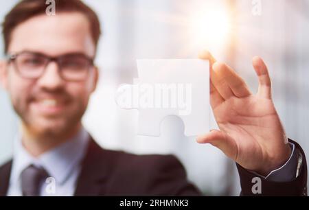 Portrait d'un homme d'affaires optimiste tenant des pièces de puzzle, résolvant des problèmes, regardant la caméra, portant un costume noir. Banque D'Images