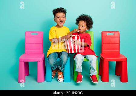 Photo grandeur nature de deux adorables petits garçons chaises assises mangeant du pop-corn porter une tenue tendance isolée sur fond de couleur aigue-marine Banque D'Images