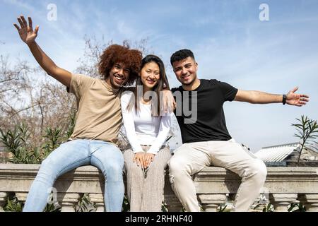 Groupe multiracial de jeunes souriants assis sur un balcon en pierre. Trois amis heureux divers s'amusant ensemble dans un parc. Banque D'Images