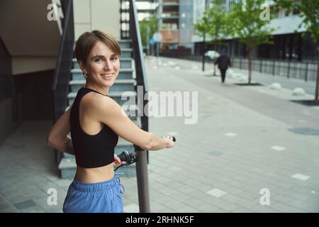 Souriant jeune femme de ville se tient avec un scooter près des escaliers, elle est en vêtements de sport confortables Banque D'Images