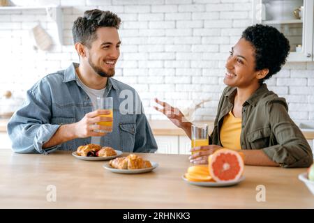 Couple heureux positif, une femme afro-américaine et un homme caucasien, bavardent assis à la maison dans la cuisine, sourire, boire du jus avec des croissants, passer du temps libre ensemble Banque D'Images
