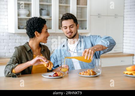 Couple heureux amoureux, une femme afro-américaine et un homme caucasien, bavardent assis à la maison dans la cuisine, sourire, boire du jus avec des croissants, passer du temps libre ensemble, temps de week-end Banque D'Images