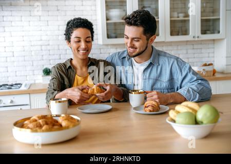 Délicieux petit déjeuner matinal. Heureux couple multiracial, homme caucasien et femme afro-américaine, passent du temps ensemble à la maison, ils boivent du café, mangent des croissants, discutent sur différents sujets, sourient Banque D'Images