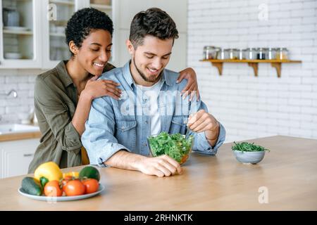 Conjoints modernes amoureux ensemble dans la cuisine, homme caucasien mange de la salade fraîche, femme afro-américaine embrasse son mari, ils passent du temps ensemble, sourient. Manger sainement, prendre soin de la santé Banque D'Images