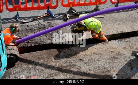 Ouvriers installant des conduits pour câble à fibre optique dans une tranchée coupée sur route avec vibreur compactant le fond de la tranchée avant que le conduit ne soit en place, Royaume-Uni Banque D'Images