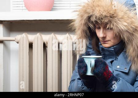 Modèle féminin moyen âge en parka et mitaines tiennent la tasse de café assis sur le sol sous le rebord de la fenêtre Banque D'Images