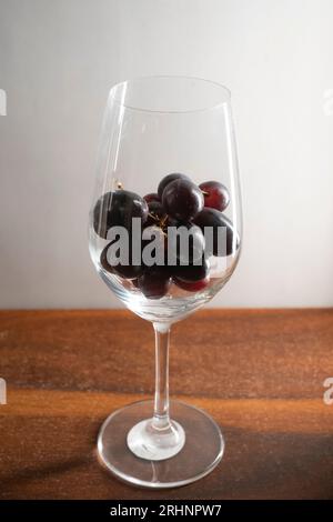 Un bouquet de raisins rouge foncé dans un verre à vin maintenu sur une table en bois sur fond blanc Banque D'Images