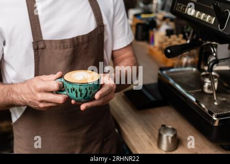 vue rognée du barista en tablier tenant la tasse de cappuccino tout en travaillant dans le café Banque D'Images