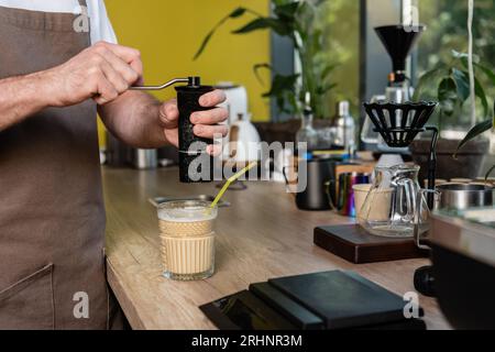 vue rognée du barista en tablier moudant du café près du café froid boire dans le verre dans le café Banque D'Images