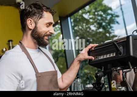 vue latérale du barista barbu et souriant dans le tablier en utilisant la machine à café tout en travaillant dans le café Banque D'Images