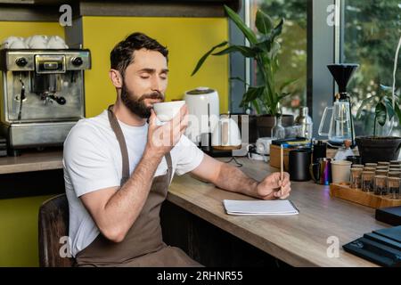 barista barbu en tablier sentant le café dans la tasse près du cahier sur l'atelier dans le café Banque D'Images