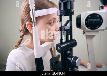 Mignonne petite fille subit un examen ophtalmique sur un équipement spécial, un enfant dans des vêtements décontractés Banque D'Images