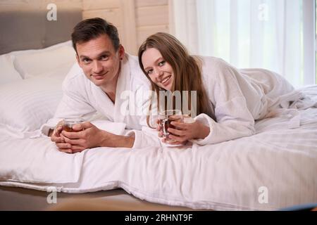 Couple souriant portant des peignoirs et couché dans le lit avec des tasses à thé Banque D'Images