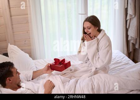 Heureuse dame donnant boîte-cadeau à l'homme souriant couché dans le lit Banque D'Images
